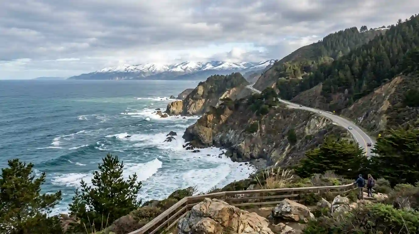 Rugged Pacific coastline with evergreen trees, winter clouds, and distant snow-covered mountains.